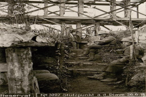 Eine Gruppe deutscher Soldaten in Helmen sitzt in einer Grube während des Zweiten Weltkriegs, umgeben von Holzpfählen und Pflanzen, mit Text am unteren Rand des Schwarz-Weiß-Bildes.
