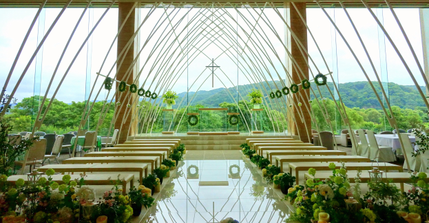 Eine Trauungszeremonie in einem großen Saal mit Tischen und Stühlen in einem Halbkreis, geschmückt mit Blumenbouquets und anderen Gegenständen, vor einer Kulisse aus Bäumen, Hügeln und einem klaren blauen Himmel.