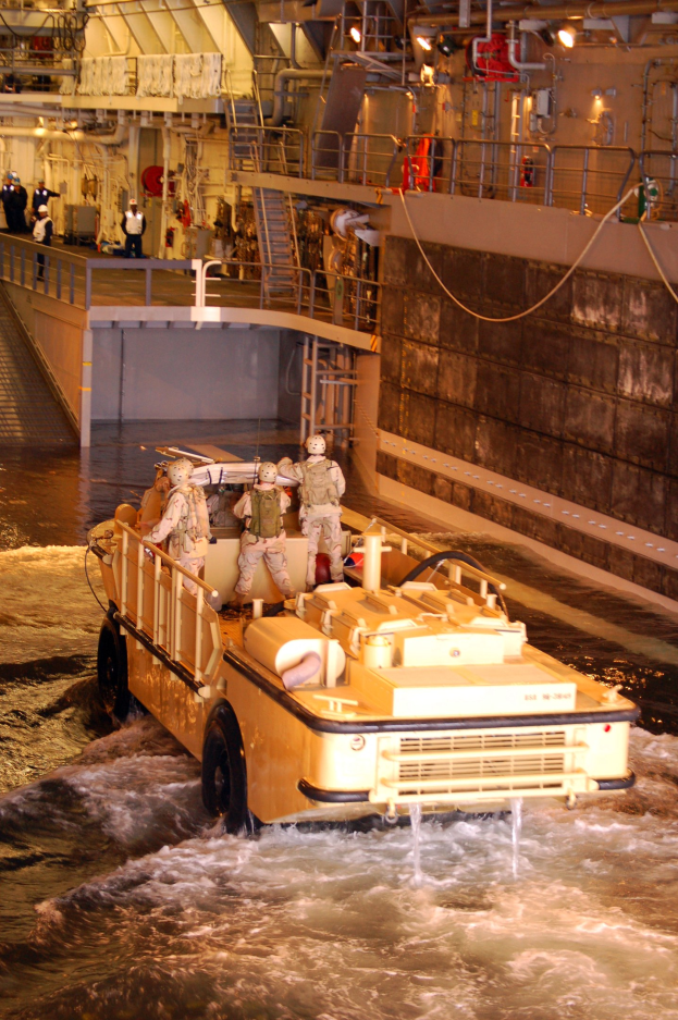 Amphibisches Fahrzeug mit Helmträgern durch Wasser in der Nähe einer Wand, mit Menschen, Geländern, Lampen und Rohren im Hintergrund.