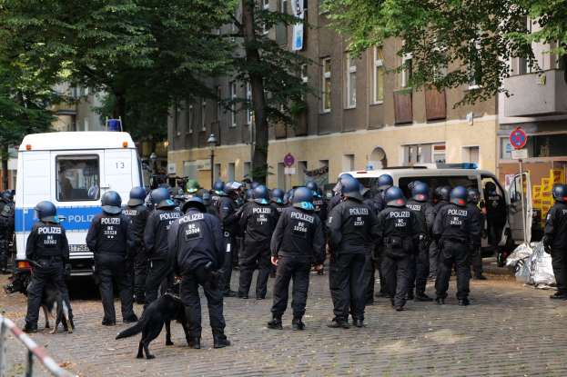 Polizeibeamte in schwarzen Uniformen und Helmen stehen vor einer Menschenmenge, mit einem schwarzen Hund im Vordergrund, Fahrzeugen und Gebäuden im Hintergrund und Bäumen, die den Bereich säumen.