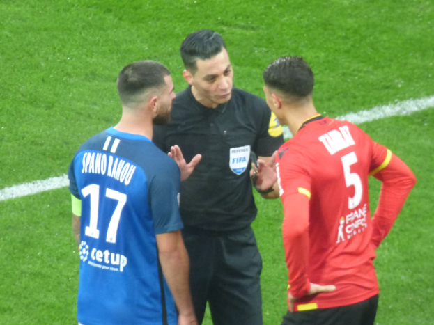 Schiedsrichter spricht mit zwei Männern auf einem Fußballfeld, einer trägt eine schwarze Hose und ein schwarzes Hemd, der andere ein blau-rotes T-Shirt mit Text, beide tragen eine Brille.