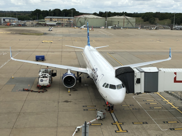 Großes Passagierflugzeug auf einem Flughafenvorfeld mit Servicefahrzeugen, Verkehrskegeln, Hallen, Polen, Bäumen und einem klaren blauen Himmel.