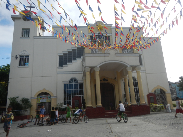Gruppe von Menschen auf Fahrrädern vor einer weißen Kirche mit architektonischen Details wie Fenstern, Säulen, Bögen, Türen und einem Kreuzsymbol, mit Pflanzen, einem Zaun, einer Fahne, Bäumen und einem bewölkten Himmel im Hintergrund und bunten Fahnen, die an einem Seil oben befestigt sind.