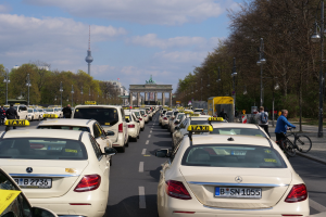Eine lange Reihe von Taxis, die an einer belebten Straße in Berlin, Deutschland, geparkt sind, mit Fahrradfahrern und Fußgängern auf dem Gehweg, flankiert von Bäumen und Laternen, und Gebäuden, einem Bogen und einem Turm im Hintergrund bei einem bewölkten Himmel.