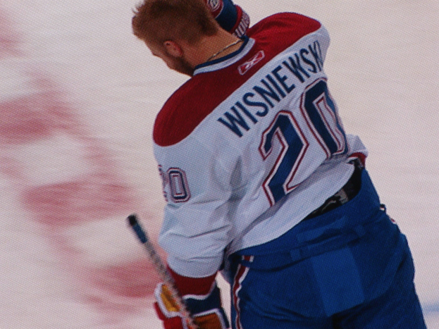 Ein Hockey-Spieler in einem "Wisniewski"-Trikot steht auf dem Eis und hält einen Hockey-Schläger in der Hand, mit einem konzentrierten und entschlossenen Gesichtsausdruck.