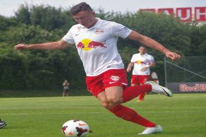 Ein Mann in einem Fußballtrikot kickt einen Ball auf einem grünen Feld mit Bäumen und einem klaren blauen Himmel im Hintergrund, mit einem Schild mit der Aufschrift "RB Leipzig" im Hintergrund.