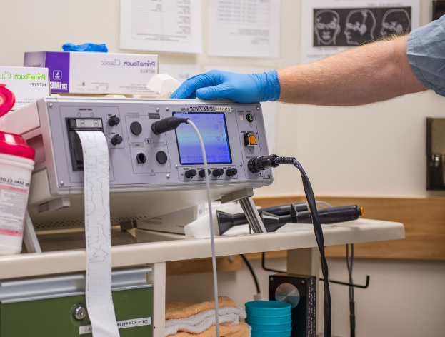 Eine Person mit blauen Handschuhen arbeitet an einem medizinischen Gerät an einem unordentlichen Tisch in einer Krankenhausumgebung mit Papieren an der Wand im Hintergrund.