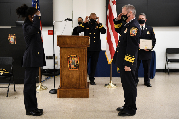 Gruppe uniformierter Personen an einem Rednerpult während einer Vereidigung eines neuen Feuerchefs, der eine Urkunde hält, umgeben von Fahnen, Stühlen, Tischen und einer Wand.