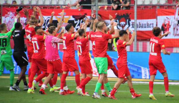 Gruppe von Fußballspielern, die ein Tor feiern, auf einem Feld mit Flaschen haltenden und klatschenden Spielern, einem Zaun, Bannern, Metallstangen, Stühlen und einer Decke mit Deckenleuchten im Hintergrund.