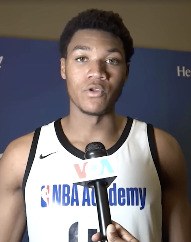 Ein Mann in einem weißen Tanktop hält ein Mikrofon und spricht mit der Presse bei der NBA All-Star-Game 2019, mit einem Banner und einer Wand im Hintergrund.