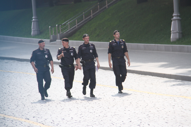 Drei Polizisten in Uniform und Mützen gehen eine Straße entlang mit Pfählen, Geländern, Treppen und Gras im Hintergrund.