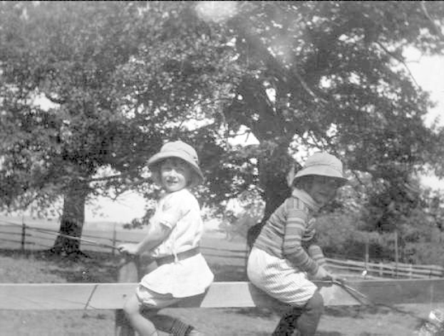 Zwei junge Jungs in Hüten halten Baseballschläger in einem grasbewachsenen Park mit Bäumen und einem Zaun im Hintergrund, das Bild in Schwarz-Weiß.