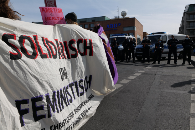 Eine Gruppe von Menschen marschiert auf einer Straße, wobei sie ein Banner mit der Aufschrift "Solidarität und Feminismus" tragen, mit parkenden Fahrzeugen und Gebäuden im Hintergrund unter einem klaren blauen Himmel.