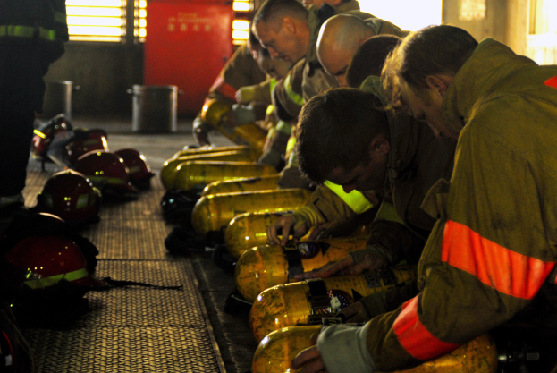 Feuerwehrleute in Schutzausrüstung bei der Arbeit an einem Feuerwehrauto mit Equipment und Helmen auf dem Boden, in der Nähe eines Gebäudes mit Fenstern und einer Tür und einem Müllcontainer.