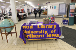 Eine Ausstellung mit einem Tisch, auf dem verschiedene Landesflaggen, eine Box, Papiere und ein Banner liegen, eine Kugel zur Linken, eine Mitteilungstafel zur Rechten mit einigen Menschen in der Nähe und mehreren Tischen und Stühlen im Hintergrund.
