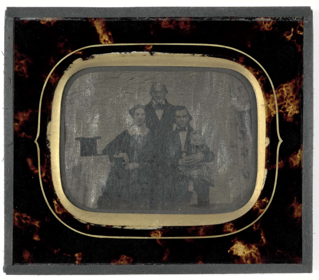 Schwarz-weißes Foto eines formell gekleideten Mannes in Anzug und Krawatte und einer Frau in einem unbekannten Outfit, die vor einem alten Fernseher stehen, beide mit ernsten Gesichtern.