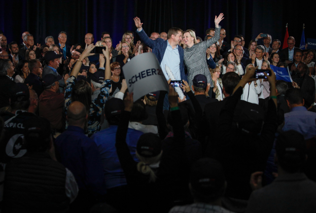 Ein Mann und eine Frau stehen vor einer Menge bei einer Wahlveranstaltung, einige halten Schilder und Mobiltelefone, sowie Fahnen und einen Vorhang im Hintergrund.
