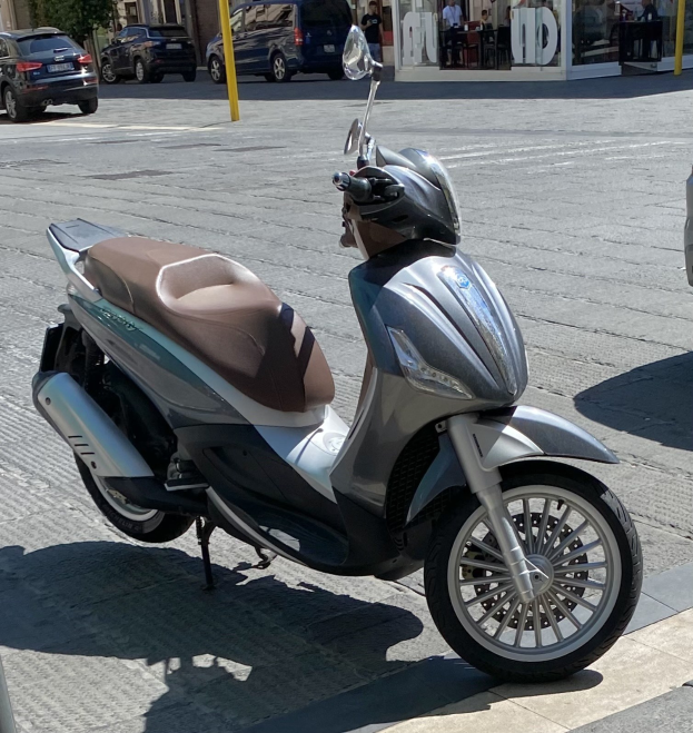 Ein Motorroller mit einem Helm darauf auf einer Straße neben Gebäuden, Bäumen und Menschen an Tischen sitzend geparkt.