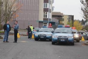 Autos auf einer Straße mit vier Menschen in der Nähe, Gebäude mit Fenstern im Hintergrund, Bäume und ein Warn Dreieck.