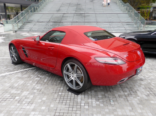Ein roter Mercedes-Benz SLS AMG Roadster vor einem Gebäude geparkt, umgeben von Menschen, Treppen, Geländern, Bäumen und anderen Gegenständen.
