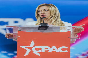 Eine Frau in einem weißen Kleid steht an einem Rednerpult mit einem Mikrofon, wobei der Hintergrund unscharf ist.