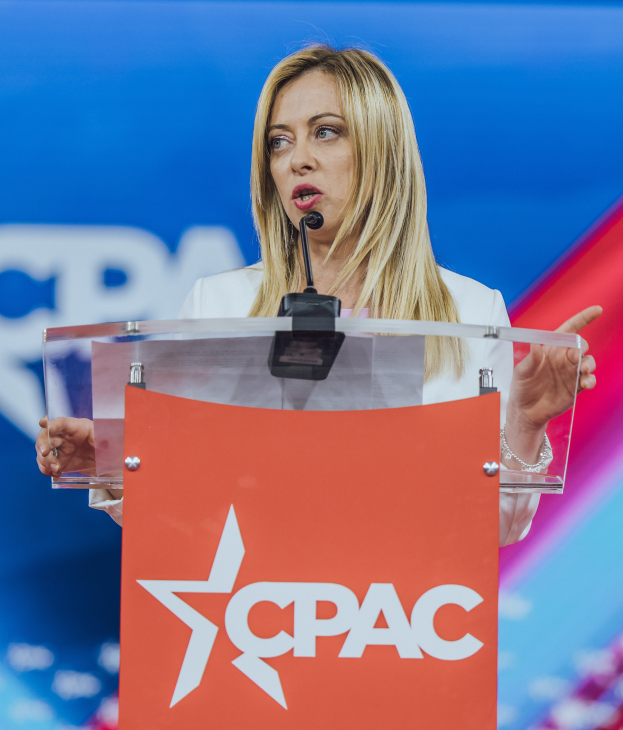 Eine Frau in einem weißen Kleid steht an einem Rednerpult mit einem Mikrofon, wobei der Hintergrund unscharf ist.