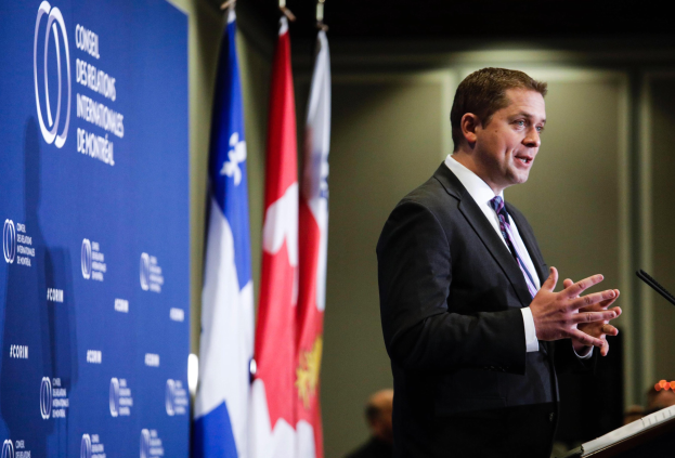 Ein Mann in Anzug und Krawatte steht an einem Rednerpult mit einem Mikrofon und spricht zu einem Publikum, mit kanadischen Flaggen und einem Banner mit der Aufschrift "Premierminister" dahinter.