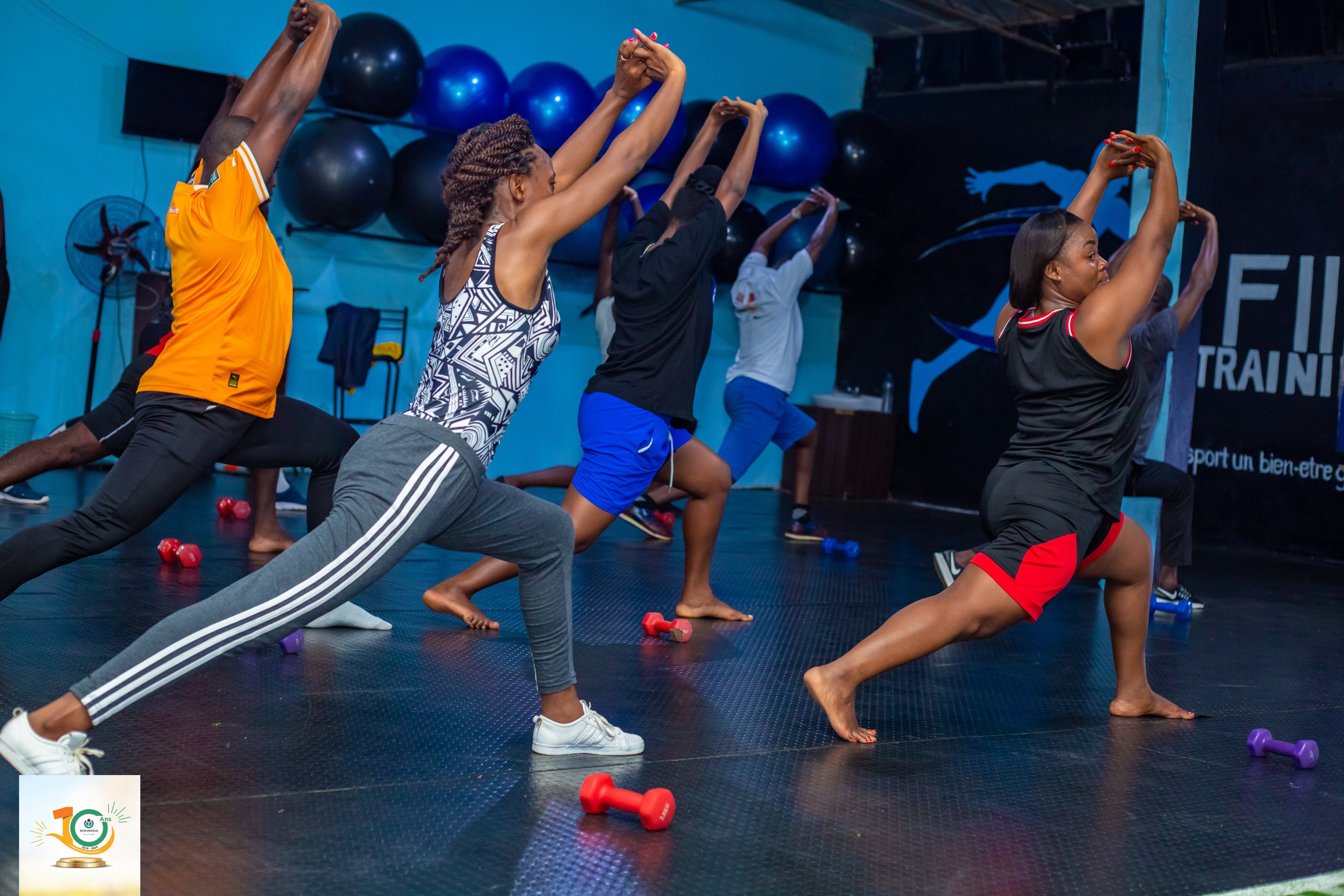 Gruppe von Menschen, die Yoga in einem Fitnessstudio mit Hanteln auf dem Boden machen, mit einem Banner "Fitness-Training in Ghana" im Hintergrund und diversen Gegenständen wie Ballons und einem Ventilator.
