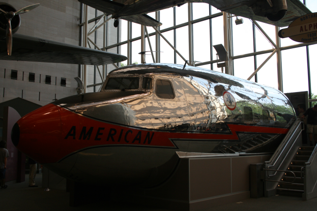 Ein Flugzeug von American Airlines in einem Museum mit Menschen drumherum, Treppe auf der rechten Seite und einem Gebäude, Bäumen und einem klaren blauen Himmel im Hintergrund.