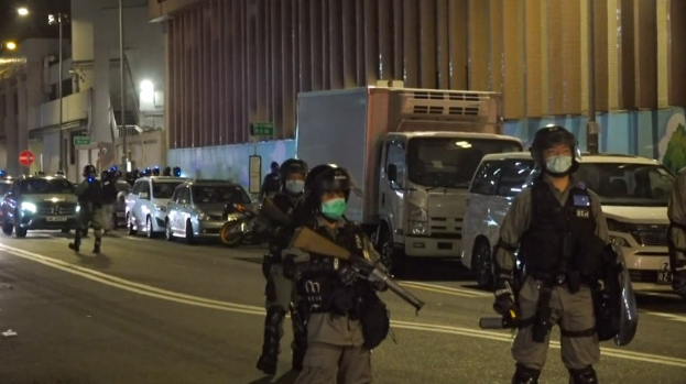 Polizeibeamte mit Helmen, Handschuhen und Gesichtsmasken stehen nachts auf einer Straße, einige halten Waffen, mit Fahrzeugen, Laternen, Schildern und Gebäuden im Hintergrund.