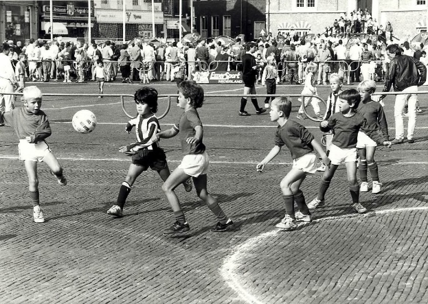 Schwarzes und weißes Bild von jungen Jungs, die Fussball auf der Straße spielen, umgeben von einer Zuschauermenge hinter einem Geländer und Gebäuden im Hintergrund.