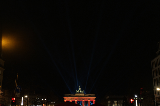Brandenburger Tor bei Nacht, beleuchtet von Straßenlaternen, mit Gebäuden, Bäumen, Pfählen, Verkehrszeichen, Schildern und Zelten im Hintergrund, vor dunklem Himmel.