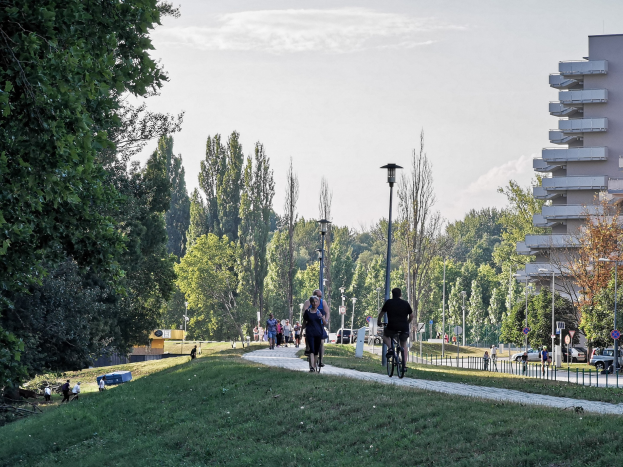 Eine Gruppe von Menschen, die auf Fahrrädern eine Parkweg entlangfahren, umgeben von Bäumen, Straßenlaternen und Gebäuden unter einem bewölkten Himmel.