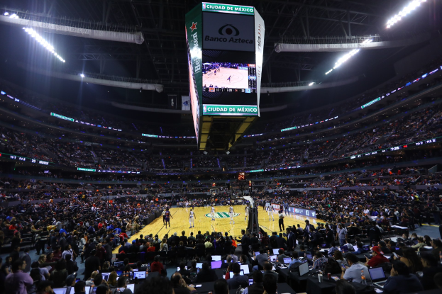Ein Basketballspiel in einer großen Arena mit Zuschauern auf Sitzplätzen und Stehplätzen, Tischen mit Laptops und Flaschen und ein Basketballkorb mit einem Anzeigebildschirm und Deckenlampen im Hintergrund.