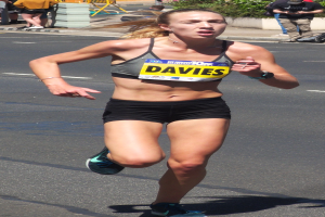 Eine Frau läuft bei einem Marathon in einem Sport-BH und Shorts, umgeben von Zuschauern auf einem Fußweg mit Pflanzen, einem Zaun, einem Schild und einem Gebäude im Hintergrund.