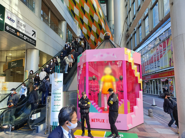 Eine Gruppe von Menschen steht vor einem Gebäude mit Glaswänden, Säulen und Texttafeln, einige tragen Masken, während eine Rolltreppe mit Menschen zu sehen ist; ein pinker Kasten in der Mitte zeigt Schaufensterpuppen und trägt die Aufschrift "Netflix's new Netflix booth in Tokyo."