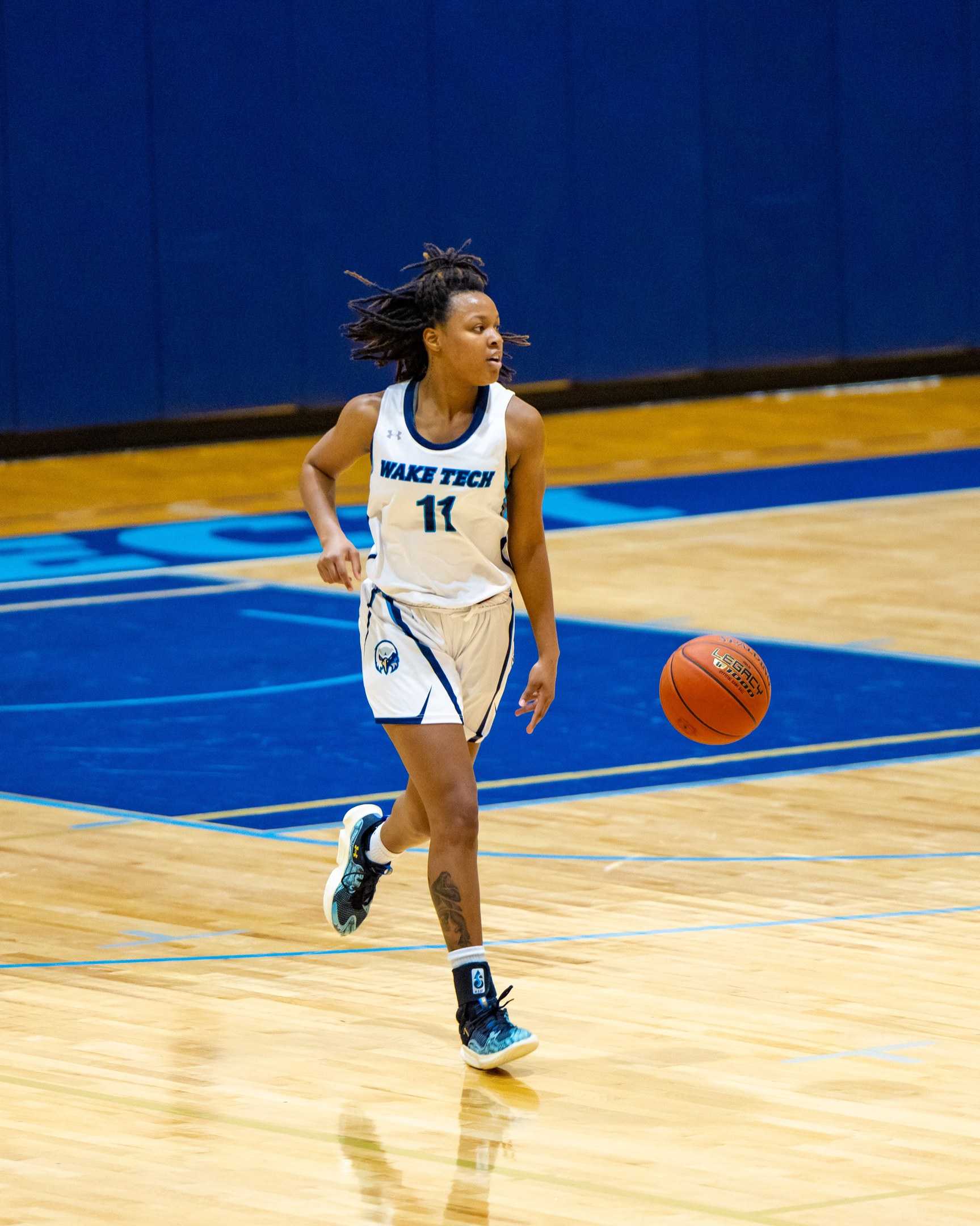 Eine Frau in einer blauen und weißen Uniform, die einen Basketball auf einem Court dribbelt, trägt ein weißes T-Shirt mit der Aufschrift "Wake Tech Women's Basketball" und blaue Schuhe, mit einer blauen Wand im Hintergrund.