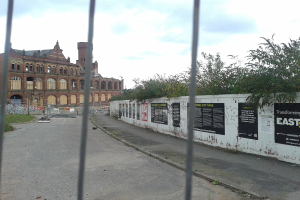 Ein großes Gebäude auf der linken Seite einer Straße, mit einer Wand und Bäumen auf der rechten Seite.