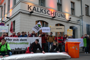 Eine Gruppe von Menschen steht vor einem Gebäude und hält Schilder und Plakate in die Höhe, während im Vordergrund zwei Personen sitzen und ein Müllcontainer rechts daneben steht, bei einer Demonstration in Deutschland, mit Gebäuden, Fenstern, Lichtern und Schildern im Hintergrund.
