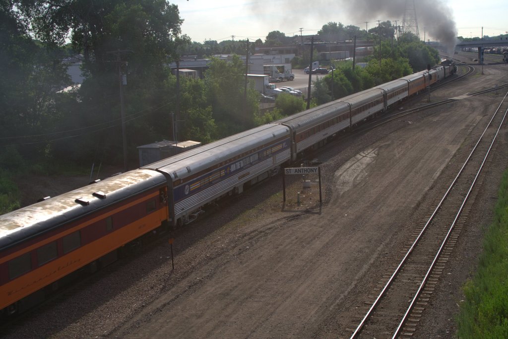 Ein Zug auf Schienen mit Bäumen, Strommasten mit Kabeln, Autos und Gebäuden links und einer Brücke vorne, aus der Rauch aufsteigt.