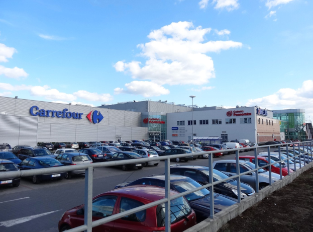 Großes Autohaus mit zahlreichen parkenden Fahrzeugen, Straßeninfrastruktur und umliegenden Gebäuden unter einem bewölkten Himmel.