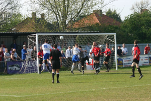 Fußballspieler spielen auf einem Feld mit einem Tor hinter ihnen, während Zuschauer von der Seitenlinie zusehen, mit Bäumen und Häusern im Hintergrund.