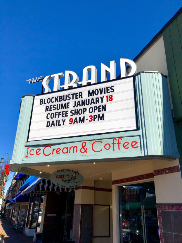 Der Strand-Blockbuster-Movies-Start am 18. Januar Kaffee-Shop-Gebäude mit einem Namensschild, einem Straßenschild, einer Gruppe von Bäumen, einigen Topfpflanzen und einem bewölkten Himmel.