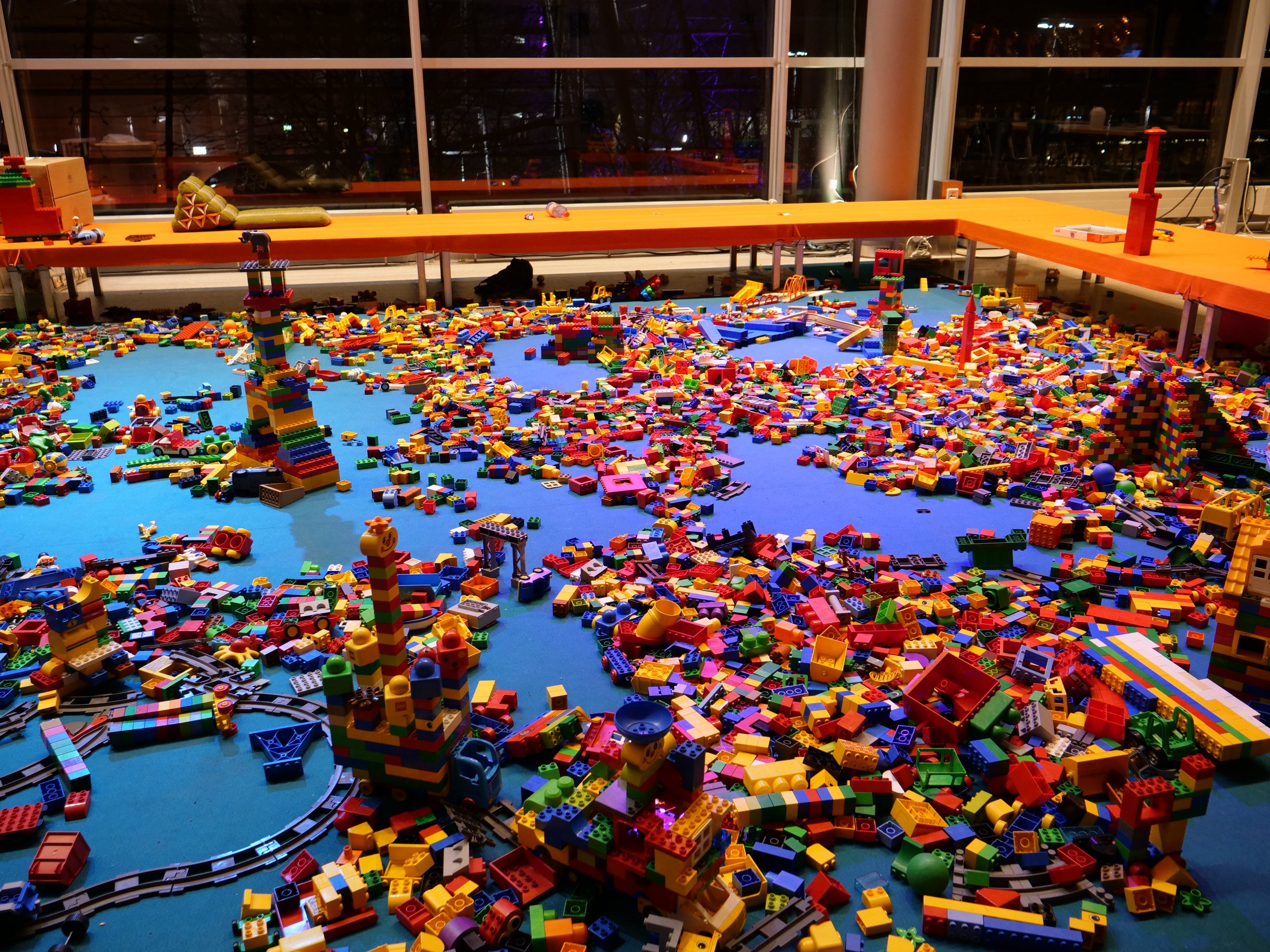 Ein großer Raum mit farbigen Lego-Steinen, die auf dem Boden verstreut sind, umgeben von Tischen und Säulen, mit Glasfenstern im Hintergrund, die Menschen und Lichter zeigen.