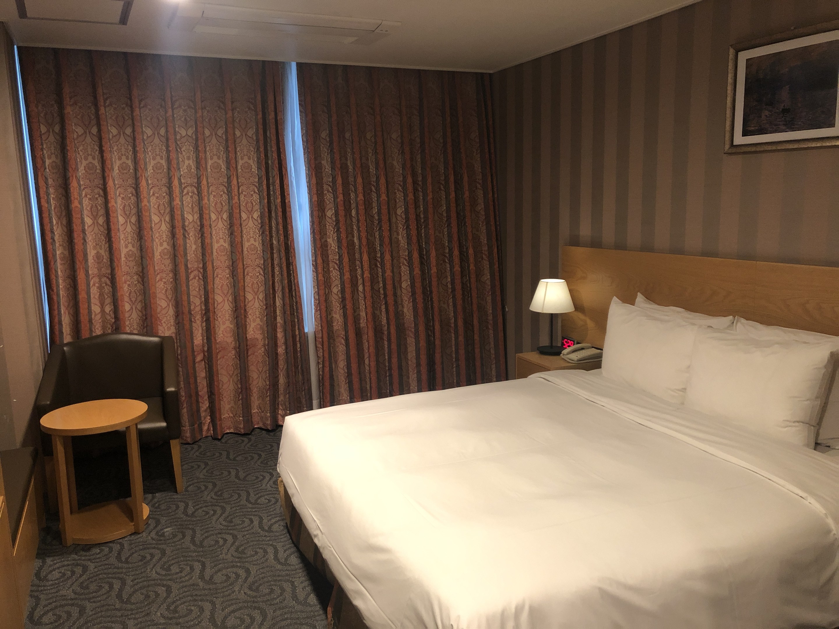 Hotelzimmer mit einem großen Bett, Stuhl, Tisch, Telefon, Lampe, Foto auf dem Wand, Fenster mit Vorhängen und Deckenbeleuchtung.
