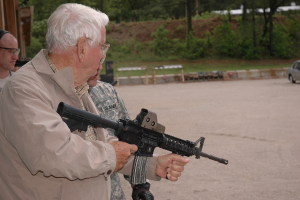 Eine Gruppe von Menschen, darunter ein Mann mit einer Waffe, steht neben einem Auto auf dem Boden mit Bäumen und Pflanzen im Hintergrund.