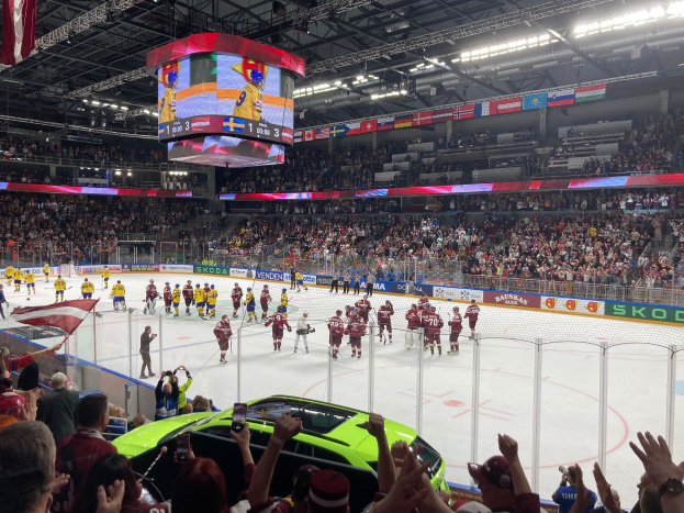 Ein Eishockeyspiel in einer großen Arena mit Zuschauern, einige halten Fahnen, ein Auto in der Nähe, eine von einem Zaun umgebene Eisfläche, Banner mit Text, Deckenleuchten und hängende Fahnen.