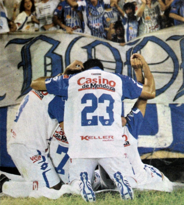 Gruppe von Fußballspielern feiert auf dem Boden vor einer jubelnden Menge mit einem Banner im Hintergrund.