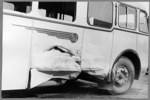 Ein Schwarz-Wei√ü-Foto eines besch√§digten Busses am Stra√üenrand, dessen Vorderseite eingedellt ist, Fenster zersplittert und die Fahrert√ľr √∂ffnet.