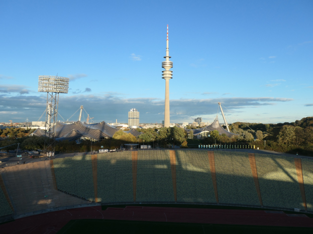 Das Olympiastadion in Berlin, Deutschland, mit dem Fernsehturm (Fernsehturm) im Hintergrund, umgeben von Bäumen, Gebäuden und beleuchteten Bereichen unter einem bewölkten Himmel.